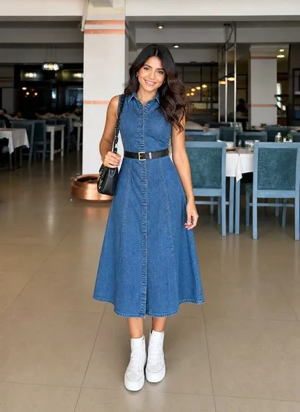 Mid-blue washed denim sleeveless shirt dress belted at the waist over a full A-line midi skirt, worn with a black quilted shoulder bag and white platform sneakers